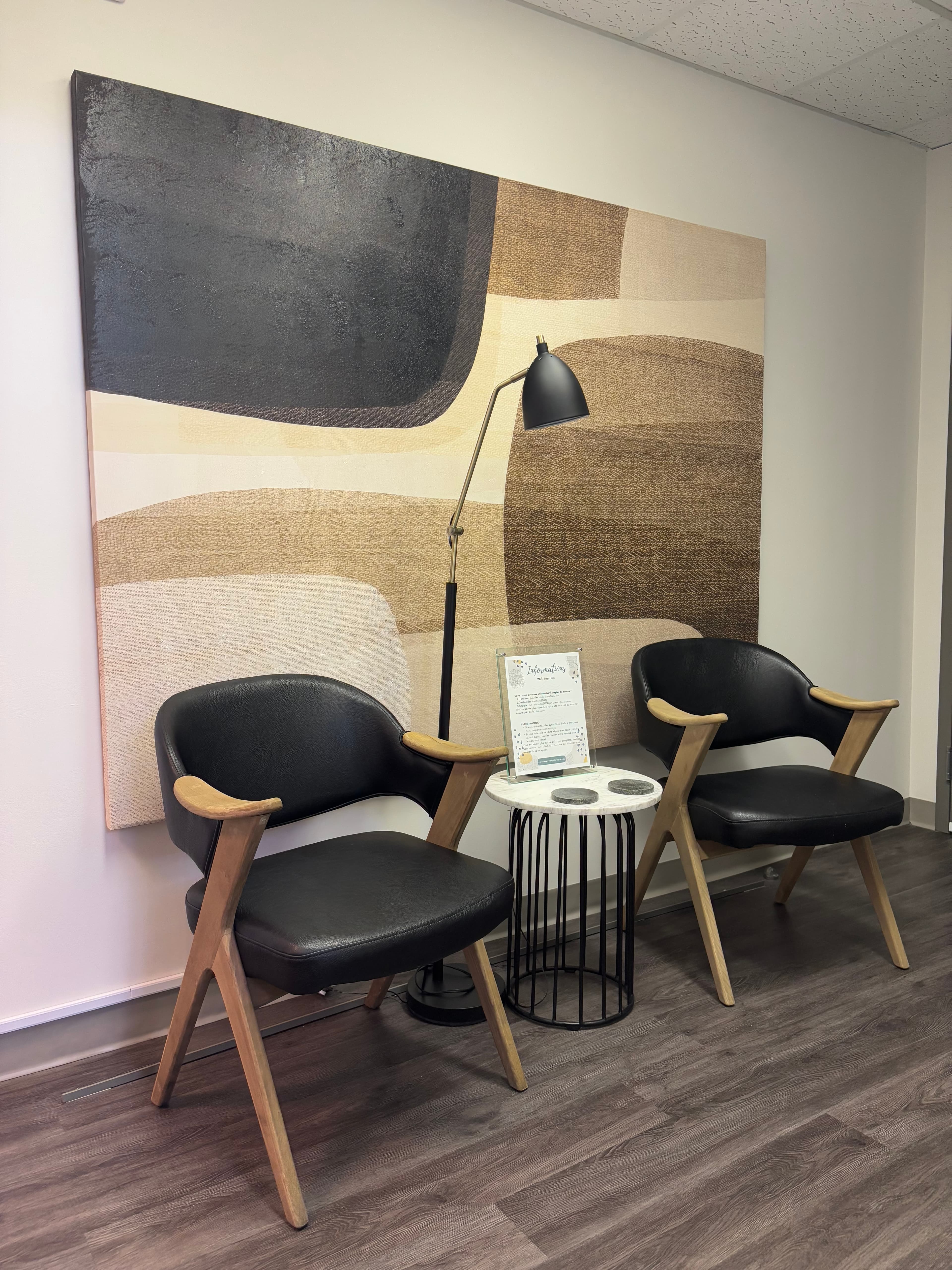 Deux fauteuils noirs et une petite table dans la salle d’attente de la clinique, devant une grande toile abstraite aux tons de beige et de brun.